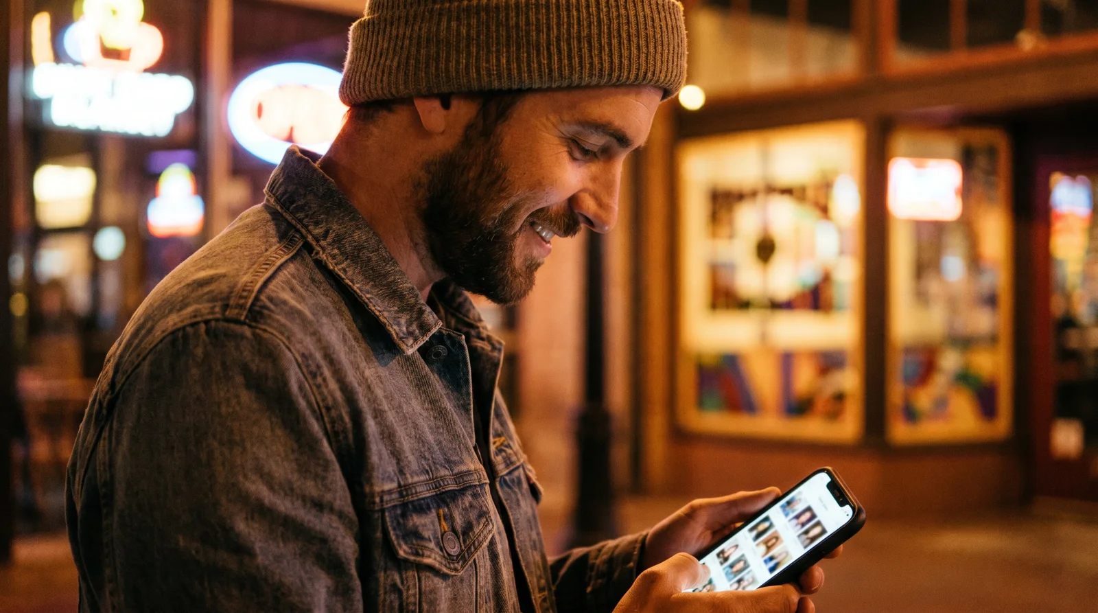 Man browsing dating profiles on a phone at night on a warm-lit Phoenix street with glowing signs behind him